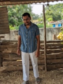 A man stands in a rustic setting, wearing a blue plaid shirt and white pants with striped sandals. The background features a wooden fence and lush greenery, suggesting an outdoor rural area.