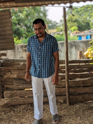 A man stands in a rustic setting, wearing a blue plaid shirt and white pants with striped sandals. The background features a wooden fence and lush greenery, suggesting an outdoor rural area.