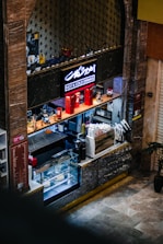 A small coffee shop counter with a display case containing pastries and cakes. The counter is equipped with a coffee machine, various bottles, and accessories for making drinks. Above, a sign with neon lighting reads 'Hot & Cold Drinks.' Decorative elements include red miniature phone booths and motorcycle models.