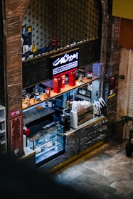 A small coffee shop counter with a display case containing pastries and cakes. The counter is equipped with a coffee machine, various bottles, and accessories for making drinks. Above, a sign with neon lighting reads 'Hot & Cold Drinks.' Decorative elements include red miniature phone booths and motorcycle models.