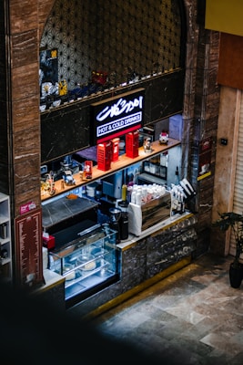 A small coffee shop counter with a display case containing pastries and cakes. The counter is equipped with a coffee machine, various bottles, and accessories for making drinks. Above, a sign with neon lighting reads 'Hot & Cold Drinks.' Decorative elements include red miniature phone booths and motorcycle models.