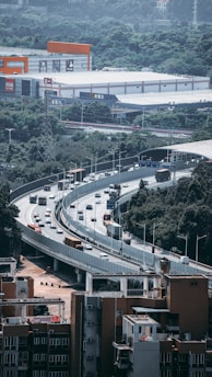 A multi-lane highway with multiple vehicles, including cars and buses, is prominently visible, elevated above a residential area. The highway curves through an urban landscape with commercial buildings and green hills in the background. There are several signs and lights along the road, contributing to the busy urban setting.