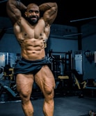 A muscular individual is posing in a gym, showcasing a highly defined and muscular physique. The lighting highlights the contours and definition of the muscles, with gym equipment visible in the background.