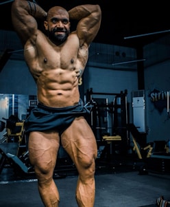 A muscular individual is posing in a gym, showcasing a highly defined and muscular physique. The lighting highlights the contours and definition of the muscles, with gym equipment visible in the background.