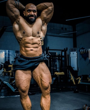A muscular individual is posing in a gym, showcasing a highly defined and muscular physique. The lighting highlights the contours and definition of the muscles, with gym equipment visible in the background.