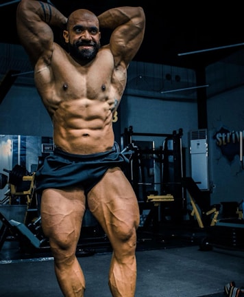 A muscular individual is posing in a gym, showcasing a highly defined and muscular physique. The lighting highlights the contours and definition of the muscles, with gym equipment visible in the background.
