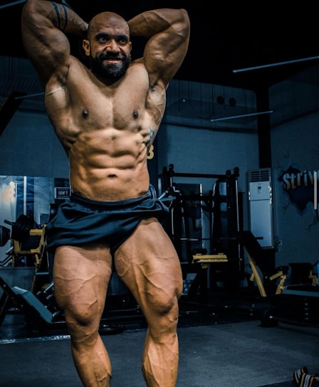 A muscular individual is posing in a gym, showcasing a highly defined and muscular physique. The lighting highlights the contours and definition of the muscles, with gym equipment visible in the background.