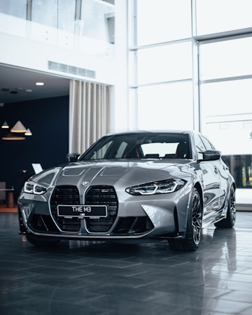 A sleek, silver BMW M3 car is prominently displayed in a modern, well-lit showroom. The car's distinct front grille and aerodynamic design are highlighted, with reflective surfaces catching the ambient light. Large windows in the background allow natural light to flood the room, casting subtle reflections on the polished floor.