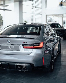 A sleek silver BMW M3 is showcased inside a well-lit showroom. The car is positioned at an angle highlighting its rear design, including the distinctive taillights and quad exhausts. Reflections on the car's surface emphasize its polished exterior.