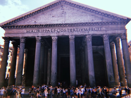 The image features a grand historical building with massive columns and a triangular pediment, inscribed with ancient Roman text. A large crowd of people gathers at the entrance, suggesting a popular tourist attraction under a bright sky.