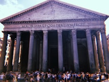 The image features a grand historical building with massive columns and a triangular pediment, inscribed with ancient Roman text. A large crowd of people gathers at the entrance, suggesting a popular tourist attraction under a bright sky.
