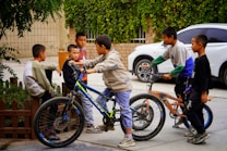 A group of young boys are gathered outside, some are sitting, while others are standing next to their bicycles. The background includes a small wooden fence, greenery, and a parked white car. The boys appear to be in conversation, possibly discussing or playing.
