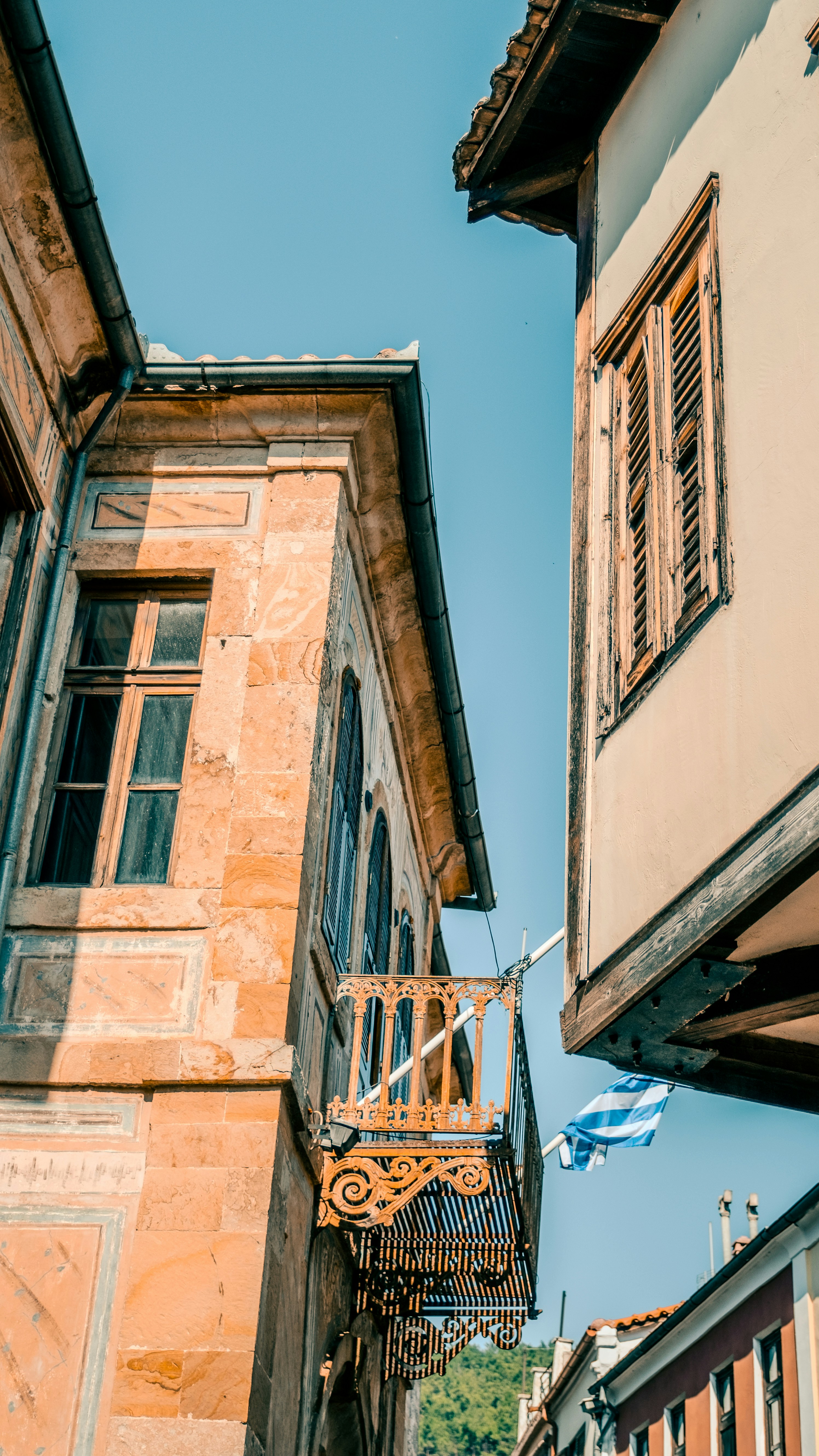 Colorful, old-style architecture featuring a small decorative balcony and wooden shutters, set against a clear blue sky. The scene is complemented by a Greek flag gently waving in the breeze.