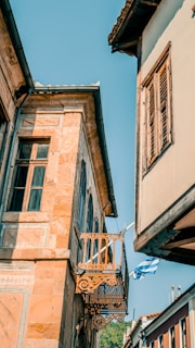 Colorful, old-style architecture featuring a small decorative balcony and wooden shutters, set against a clear blue sky. The scene is complemented by a Greek flag gently waving in the breeze.
