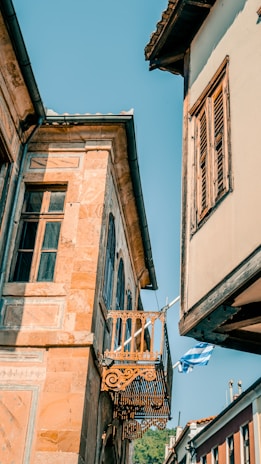 Colorful, old-style architecture featuring a small decorative balcony and wooden shutters, set against a clear blue sky. The scene is complemented by a Greek flag gently waving in the breeze.