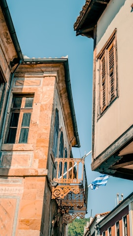 Colorful, old-style architecture featuring a small decorative balcony and wooden shutters, set against a clear blue sky. The scene is complemented by a Greek flag gently waving in the breeze.