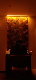 A person sits at a desk under warm, ambient lighting. The wall behind them features a geometric pattern with a backlit panel that emits a soft yellow-orange glow. On the desk, there are various items, including a white bag and a few other objects, possibly food wrappers or small items.
