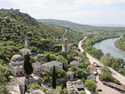 A picturesque village nestled alongside a gently curving river, surrounded by lush greenery and rolling hills. The settlement features traditional stone buildings with tiled roofs, a prominent mosque with a tall minaret, and an ancient stone tower. A winding road follows the riverbank into the distance.