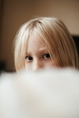 A close-up image of a young child with blonde hair partially obscured by a soft-focus object in the foreground. The child’s eyes are visible, looking directly at the camera with a neutral expression. The background is softly lit, contributing to a warm, intimate atmosphere.