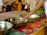 A person with a bracelet is reaching towards a collection of metal bowls placed on a table, each filled with various offerings including money. Several people stand in the background, some wearing traditional attire. The setting appears to be a ceremonial or religious occasion, as suggested by the ornate decorations and vibrant colors.