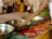 A person with a bracelet is reaching towards a collection of metal bowls placed on a table, each filled with various offerings including money. Several people stand in the background, some wearing traditional attire. The setting appears to be a ceremonial or religious occasion, as suggested by the ornate decorations and vibrant colors.