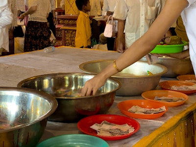 A person with a bracelet is reaching towards a collection of metal bowls placed on a table, each filled with various offerings including money. Several people stand in the background, some wearing traditional attire. The setting appears to be a ceremonial or religious occasion, as suggested by the ornate decorations and vibrant colors.