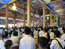 A large group of people, dressed mostly in white, are gathered inside a vividly decorated temple with elaborate wall and ceiling paintings, intricate columns, and vibrant lighting. Some monks in orange robes can be seen at the front of the room.