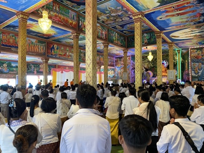A large group of people, dressed mostly in white, are gathered inside a vividly decorated temple with elaborate wall and ceiling paintings, intricate columns, and vibrant lighting. Some monks in orange robes can be seen at the front of the room.