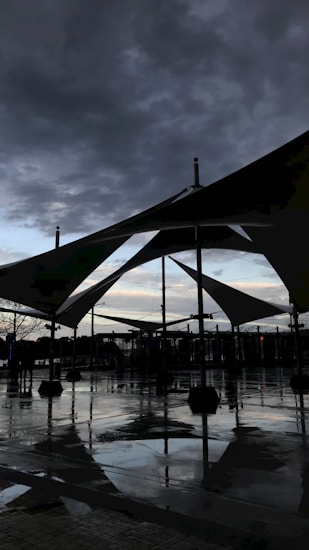 Large geometric canopy structures with triangular shapes are silhouetted against a cloudy sky. The ground is wet, reflecting the structures and dark clouds above, suggesting recent rain. A few people can be seen in the distance, and there are various lights visible, likely from street lamps or nearby buildings.