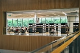 A spacious library interior with people seated at tables, engaged in reading or studying. Tall bookshelves lined with books border the open space. Large windows in the background reveal lush greenery outside, allowing ample natural light to fill the room.