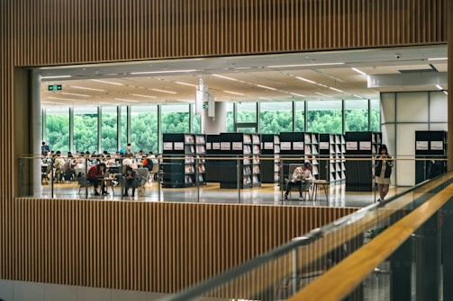 A spacious library interior with people seated at tables, engaged in reading or studying. Tall bookshelves lined with books border the open space. Large windows in the background reveal lush greenery outside, allowing ample natural light to fill the room.