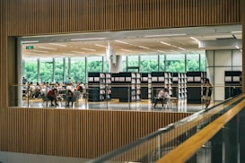 A spacious library interior with people seated at tables, engaged in reading or studying. Tall bookshelves lined with books border the open space. Large windows in the background reveal lush greenery outside, allowing ample natural light to fill the room.