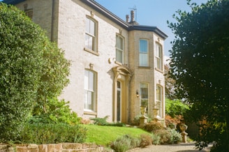 A large two-story house made of light-colored brick with several tall windows and a front door adorned with an arch. The house is surrounded by well-maintained greenery, including shrubs and trees, creating a welcoming and lush garden setting.