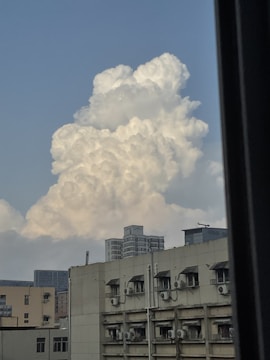 Large, fluffy cumulus clouds rise prominently against a blue sky, providing a striking contrast with a cityscape in the foreground. The buildings feature multiple windows and air conditioning units are visible on the exterior walls.