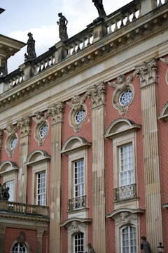 An ornate facade of a historic building features red brick with intricate stone carvings and decorative elements, including statues on the rooftop. Tall windows with white frames are accented with decorative molding and wrought iron railings.