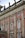 An ornate facade of a historic building features red brick with intricate stone carvings and decorative elements, including statues on the rooftop. Tall windows with white frames are accented with decorative molding and wrought iron railings.