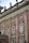 An ornate facade of a historic building features red brick with intricate stone carvings and decorative elements, including statues on the rooftop. Tall windows with white frames are accented with decorative molding and wrought iron railings.