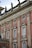 An ornate facade of a historic building features red brick with intricate stone carvings and decorative elements, including statues on the rooftop. Tall windows with white frames are accented with decorative molding and wrought iron railings.