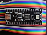 A microcontroller board is positioned on top of a series of multi-colored jumper wires. The board features various electronic components including a USB port, buttons, and several surface-mounted chips. The jumper wires are organized and display a gradient of colors including red, yellow, green, blue, purple, and white.