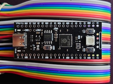 A microcontroller board is positioned on top of a series of multi-colored jumper wires. The board features various electronic components including a USB port, buttons, and several surface-mounted chips. The jumper wires are organized and display a gradient of colors including red, yellow, green, blue, purple, and white.
