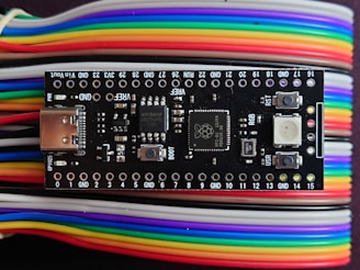 A microcontroller board is positioned on top of a series of multi-colored jumper wires. The board features various electronic components including a USB port, buttons, and several surface-mounted chips. The jumper wires are organized and display a gradient of colors including red, yellow, green, blue, purple, and white.