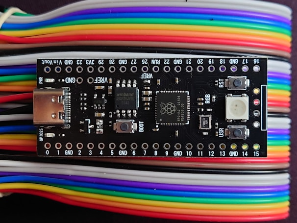A microcontroller board is positioned on top of a series of multi-colored jumper wires. The board features various electronic components including a USB port, buttons, and several surface-mounted chips. The jumper wires are organized and display a gradient of colors including red, yellow, green, blue, purple, and white.