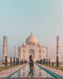 A person stands in front of the iconic Taj Mahal, with its white marble structure prominently displayed. The Taj Mahal is framed by two of its minarets, with one under restoration. A reflective pool runs in the center, bordered by manicured trees. Birds are flying in the sky above the monument.