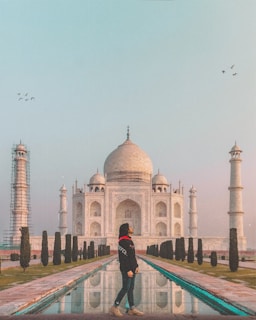 A person stands in front of the iconic Taj Mahal, with its white marble structure prominently displayed. The Taj Mahal is framed by two of its minarets, with one under restoration. A reflective pool runs in the center, bordered by manicured trees. Birds are flying in the sky above the monument.