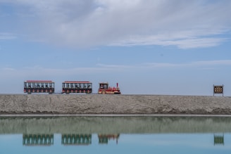 A small tourist train with two carriages and a red engine travels along a raised path, beside a reflective body of water on a clear day. A sign with text is visible on the right side.