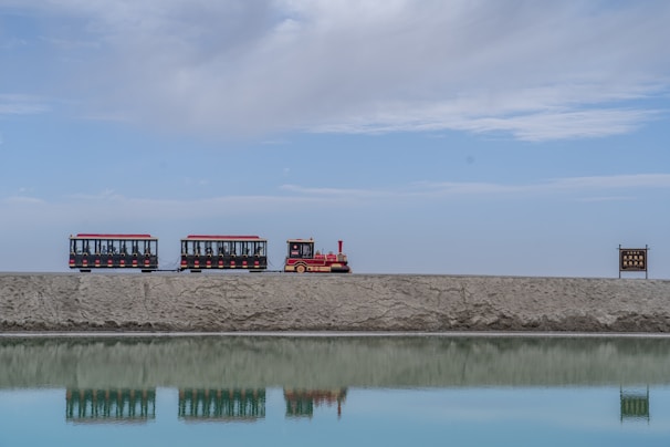 A small tourist train with two carriages and a red engine travels along a raised path, beside a reflective body of water on a clear day. A sign with text is visible on the right side.