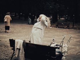 An elderly person with long white hair sits on a park bench, readjusting their hair. They are dressed in a light-colored coat. Nearby, an empty shopping cart is filled with various bags or blankets. The setting is a park with stone pathways and trees in the background. Another person is walking away in the distance.