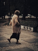 An elderly person walks along a cobblestone path in an outdoor setting. The individual is wearing a long beige coat and carrying a handbag. The surroundings include trees and a bench with graffiti on it, creating an urban park-like atmosphere.