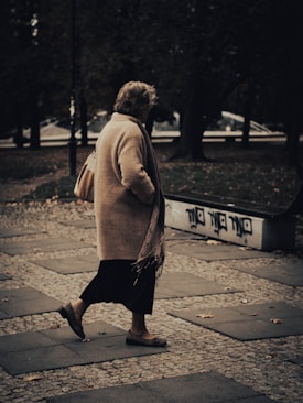 An elderly person walks along a cobblestone path in an outdoor setting. The individual is wearing a long beige coat and carrying a handbag. The surroundings include trees and a bench with graffiti on it, creating an urban park-like atmosphere.
