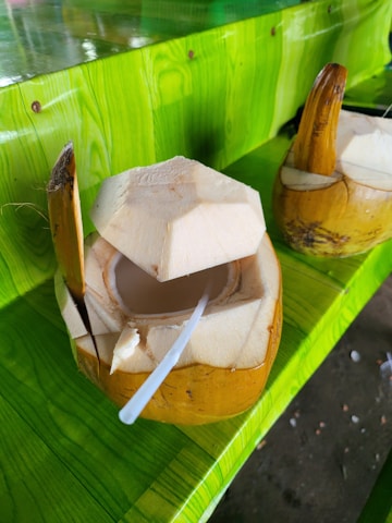 Two freshly cut coconuts are placed on a vibrant, green surface. One coconut is opened with a straw inserted, ready for drinking, while the other remains sealed. The lush green color of the table contrasts with the natural brown and cream tones of the coconuts.